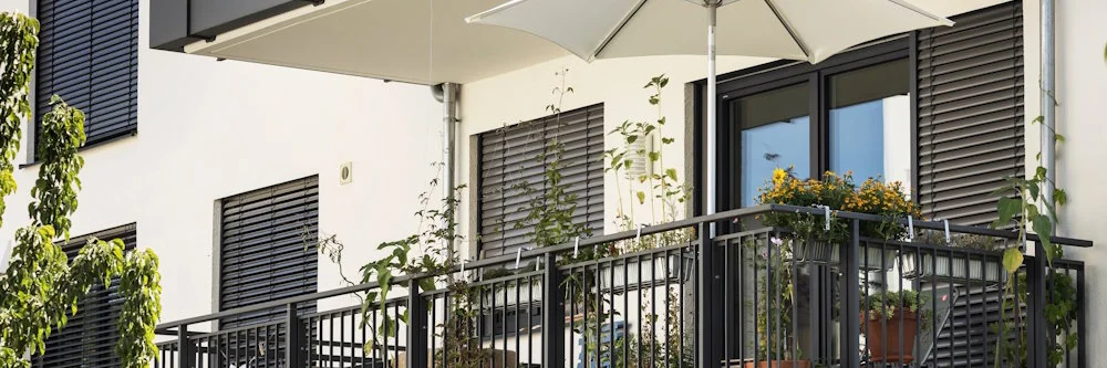 Moderner Balkon mit Sonnenschirm, Pflanzen und dunklen Jalousien.